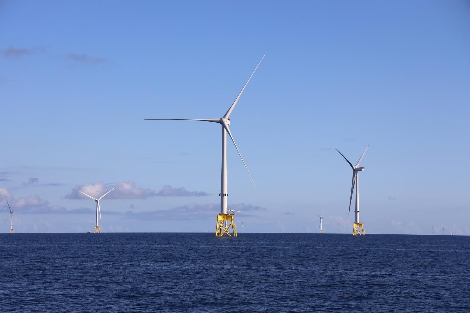 Un aerogenerador grande en primer plano y otros más pequeños hacia el horizonte, sobre bases amarillas en mar abierto bajo cielo azul